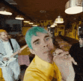 a man with green hair is singing into a microphone in a restaurant while another man plays a guitar .