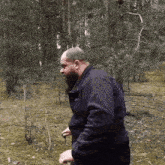 a man with a beard in a black jacket is walking through a forest
