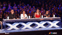 a group of judges sit at a table with a large x on it in front of a crowd