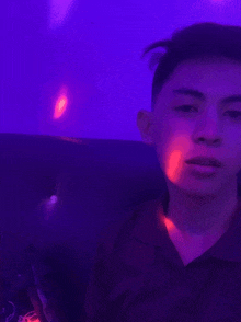 a young man is sitting on a couch in front of a purple wall holding a cell phone .