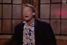 a man in a hawaiian shirt is standing in front of a window with a snl logo on the bottom right corner