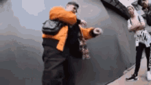 a man in an orange jacket is dancing in front of a group of people standing in front of a wall .