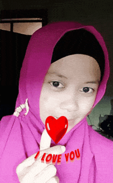 a girl wearing a pink hijab is holding a red heart with the words " i love you " written below it