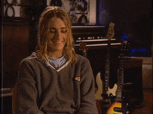 a man wearing a grey sweatshirt with a s on it smiles in front of a keyboard and guitars