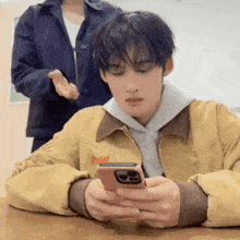 a man in a yellow jacket is sitting at a table looking at his cell phone .