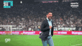 a man in a suit stands on a soccer field in front of a scoreboard that says sportv