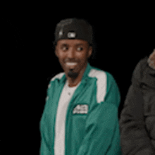 a man in a green jacket and a black hat is standing next to another man in a black jacket .