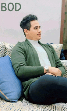 a man in a green cardigan sits on a couch with the word bod behind him