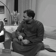 a black and white photo of a man sitting on a couch holding a rosary and a plaque with arabic writing