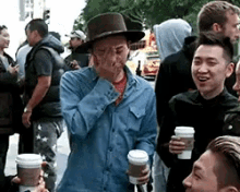 a man wearing a hat is covering his face while standing in a crowd of people holding coffee cups .
