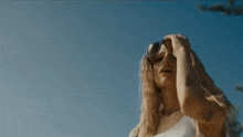 a woman with long blonde hair is standing in front of a blue sky and trees .