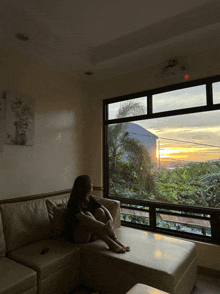 a woman sits on a couch in front of a window looking out at the sunset