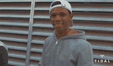 a man wearing a white hat and a gray hoodie is smiling in front of a wall with the word tidal on it