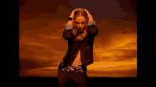 a woman in a denim jacket is dancing in front of a sunset sky