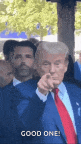 a man in a suit and tie is pointing at the camera while another man looks on .