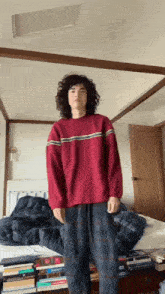 a man with curly hair wearing a red sweater and plaid pants stands in front of a bed