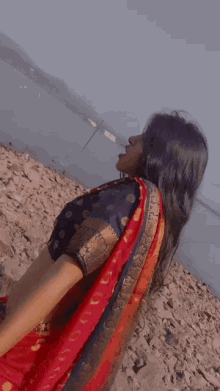 a woman in a red and gold polka dot saree stands on a rocky shore