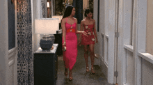 two women in pink dresses walk down a hallway holding hands