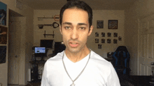 a man in a white shirt with a chain around his neck is standing in front of a wall with pictures on it