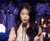 a woman in a white dress is holding a trophy in front of two microphones with the letter k on the bottom right