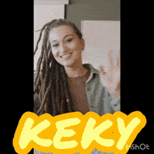 a woman with dreadlocks is smiling and waving in front of a sign that says keky