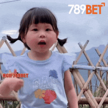 a little girl wearing a gap t-shirt stands in front of a bamboo fence
