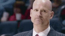 a bald man in a suit and tie is making a funny face while sitting in a stadium .