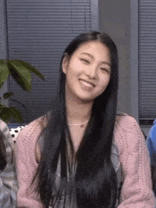 a woman with long black hair is wearing a pink sweater and smiling while sitting on a couch .
