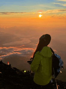 a woman in a yellow jacket stands on top of a mountain looking at the sunset