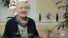 a bald man with a scarf around his neck smiles in front of a plant