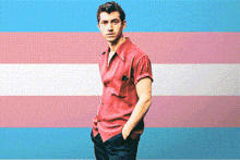 a man in a red shirt and black pants is standing in front of a transgender flag .