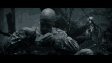 a bald man in a military uniform is covering his face with his hand in a dark forest .