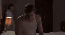 a man in a white tank top is sitting on a bed with a woman standing in the background .
