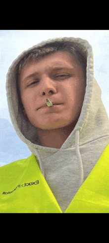 a man wearing a grey hoodie and a yellow vest with the word excel on it smoking a cigarette