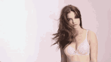 a woman in a pink bra is standing in front of a pink background with her hair blowing in the wind .