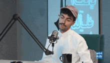 a man wearing a hat is sitting in front of a microphone with arabic writing behind him