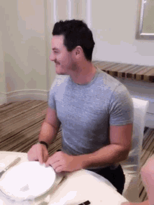 a man in a grey shirt sits at a table with plates and knives