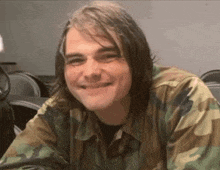 a man with long hair wearing a camouflage shirt smiles