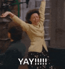 a woman in a yellow shirt is dancing in a hallway with her arms in the air and the words yay !