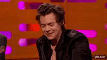 harry styles is wearing a black suit and a cross necklace while laughing on a stage .