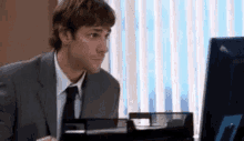 a man in a suit and tie is sitting at a desk looking at a computer screen .