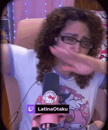 a woman wearing glasses and a hello kitty shirt is covering her mouth with her hands in front of a pink hello kitty microphone