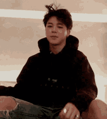 a young man is sitting on a bed wearing a black hoodie and ripped jeans .