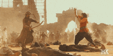 a man with a sword is standing in front of a crowd with the letters hk on the bottom right