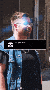 a man wearing glasses and a denim vest is standing in front of a skull that says you 're