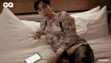 a man is laying on a bed with a phone in his hand and a gq logo in the background