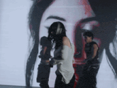 a woman in a white shirt is standing in front of a large screen with a woman 's face projected on it