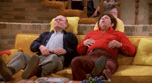 two men are sitting on a yellow couch with their heads on pillows