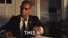 a man in a suit and tie is sitting at a desk with a book and glasses .
