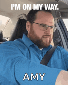 a man in a blue shirt and glasses is driving a car and says " i 'm on my way amy "